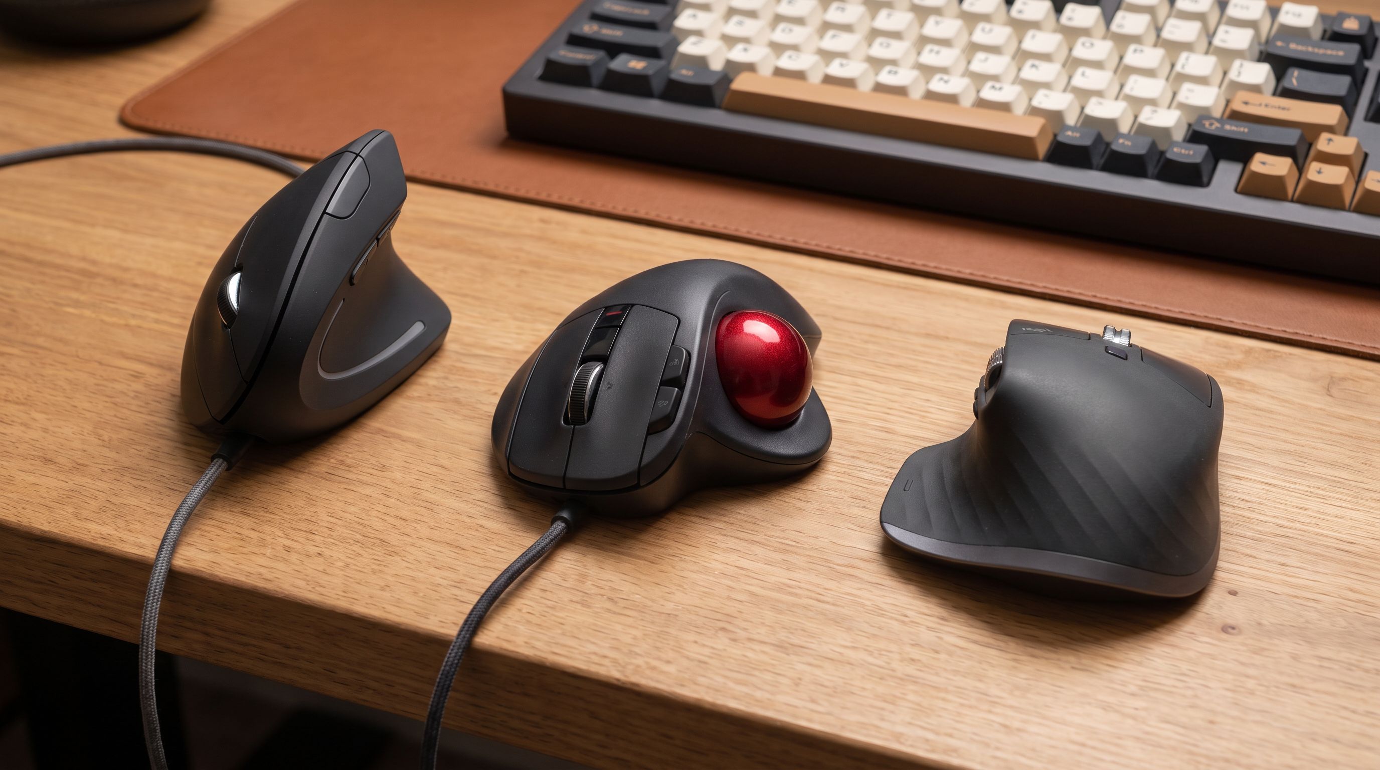 A vertical ergonomic mouse, a thumb trackball and a sculpted wireless mouse arranged on a wooden desk beside a keyboard
