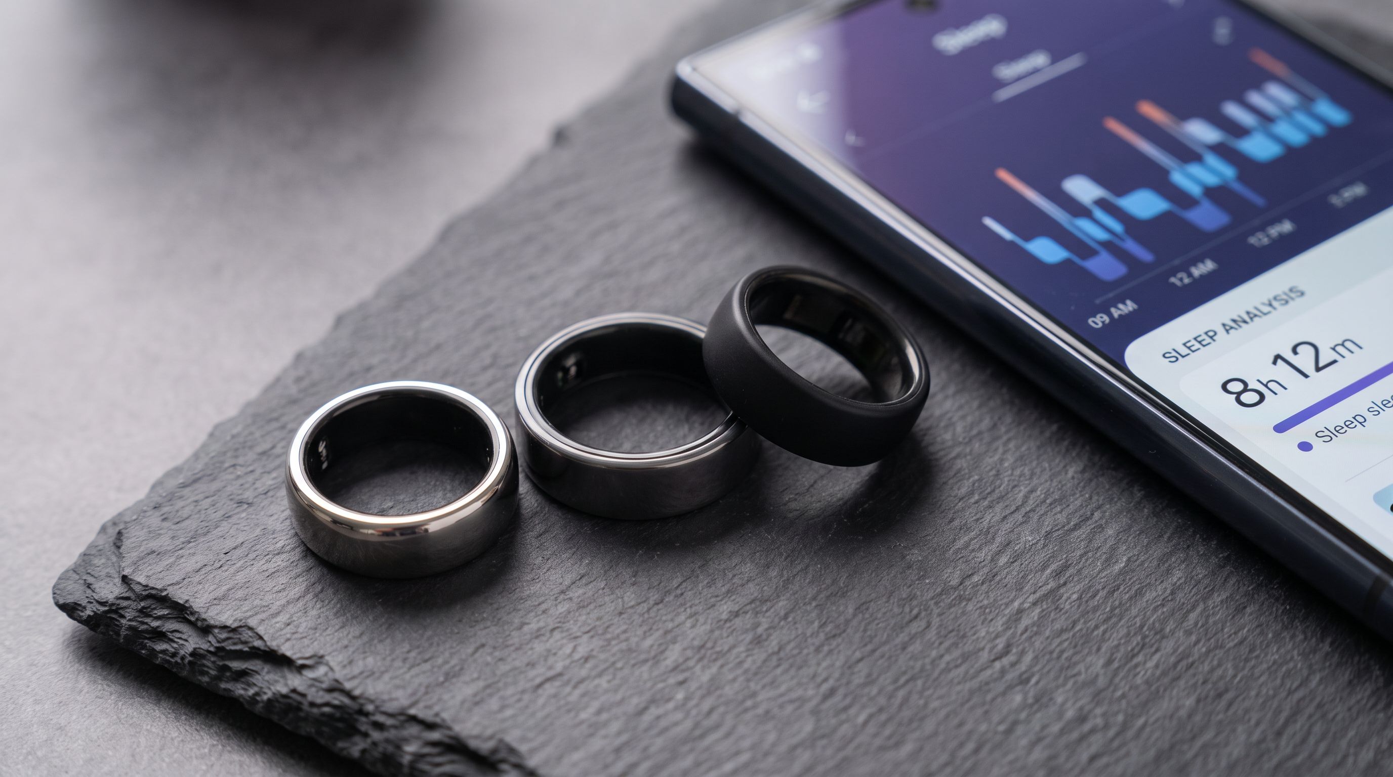 Three smart rings — a titanium Oura Ring 4, a Samsung Galaxy Ring and an Ultrahuman Ring Air — arranged on a dark surface beside a smartphone showing sleep data
