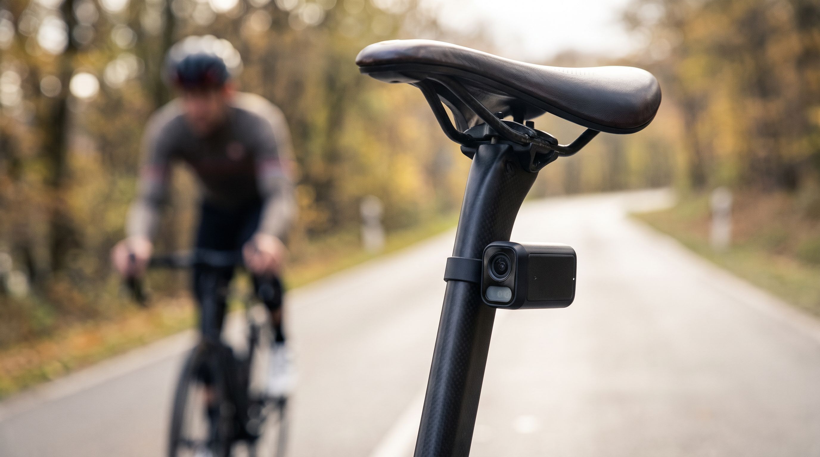 A road bike seatpost with a rear-facing radar camera mounted below the saddle, with a cyclist riding on a tree-lined road in the background