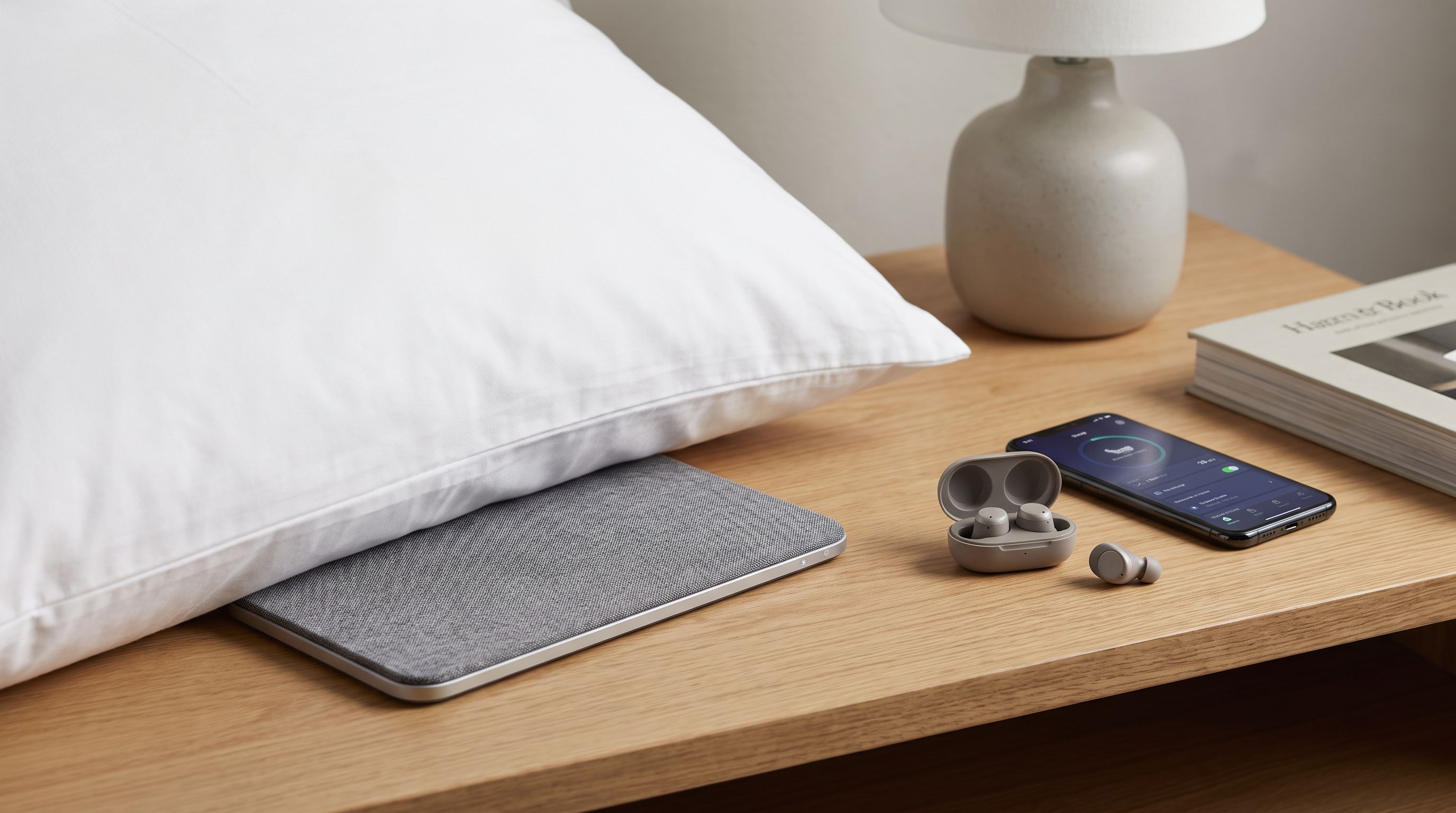 A slim Bluetooth pillow speaker tucked under a hotel pillow beside a pair of sleep earbuds and a phone on the nightstand