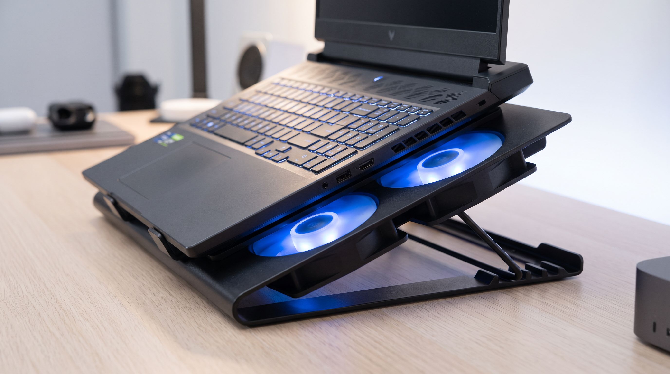 A gaming laptop sitting on a black multi-fan cooling pad with blue LED-lit fans, angled on a desk