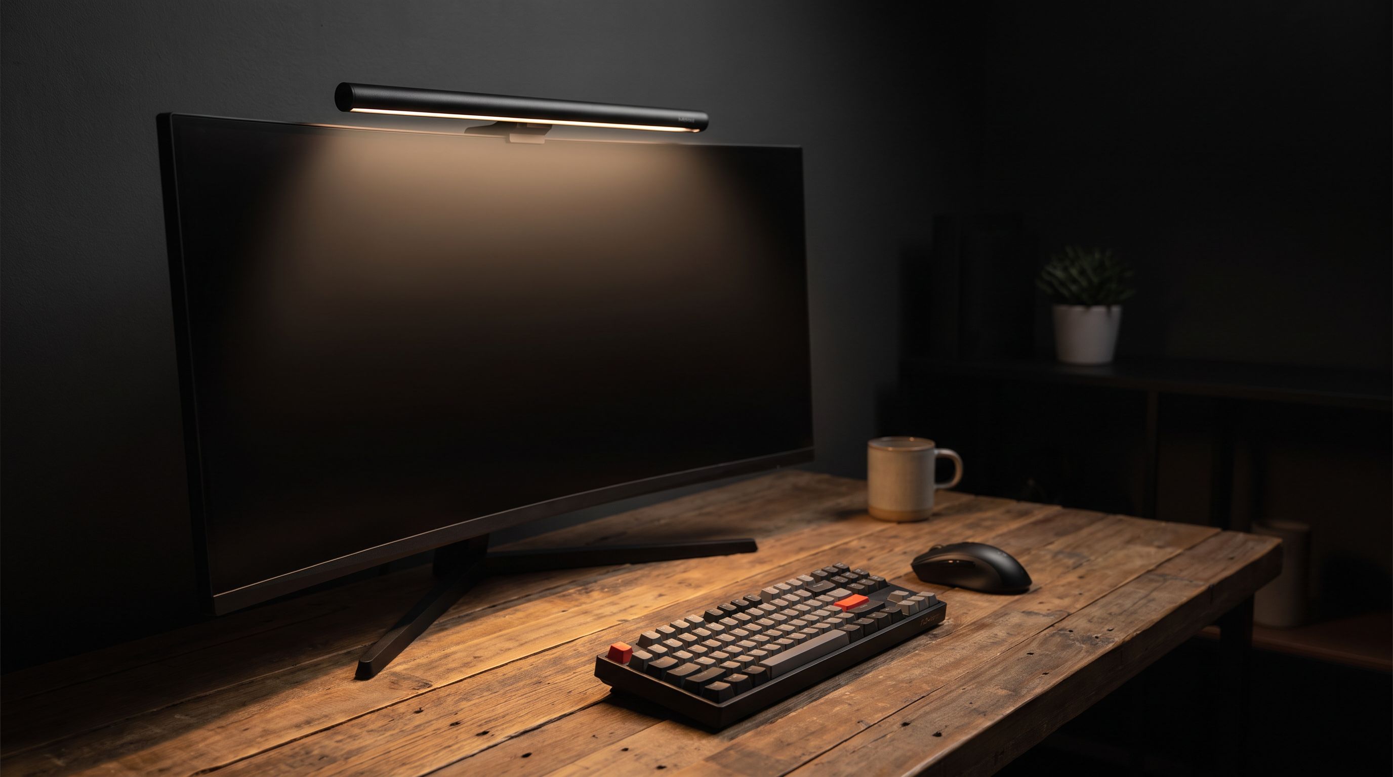 A monitor light bar clipped to the top bezel of a widescreen monitor, casting a warm even pool of light across a wooden desk and keyboard in a dim room
