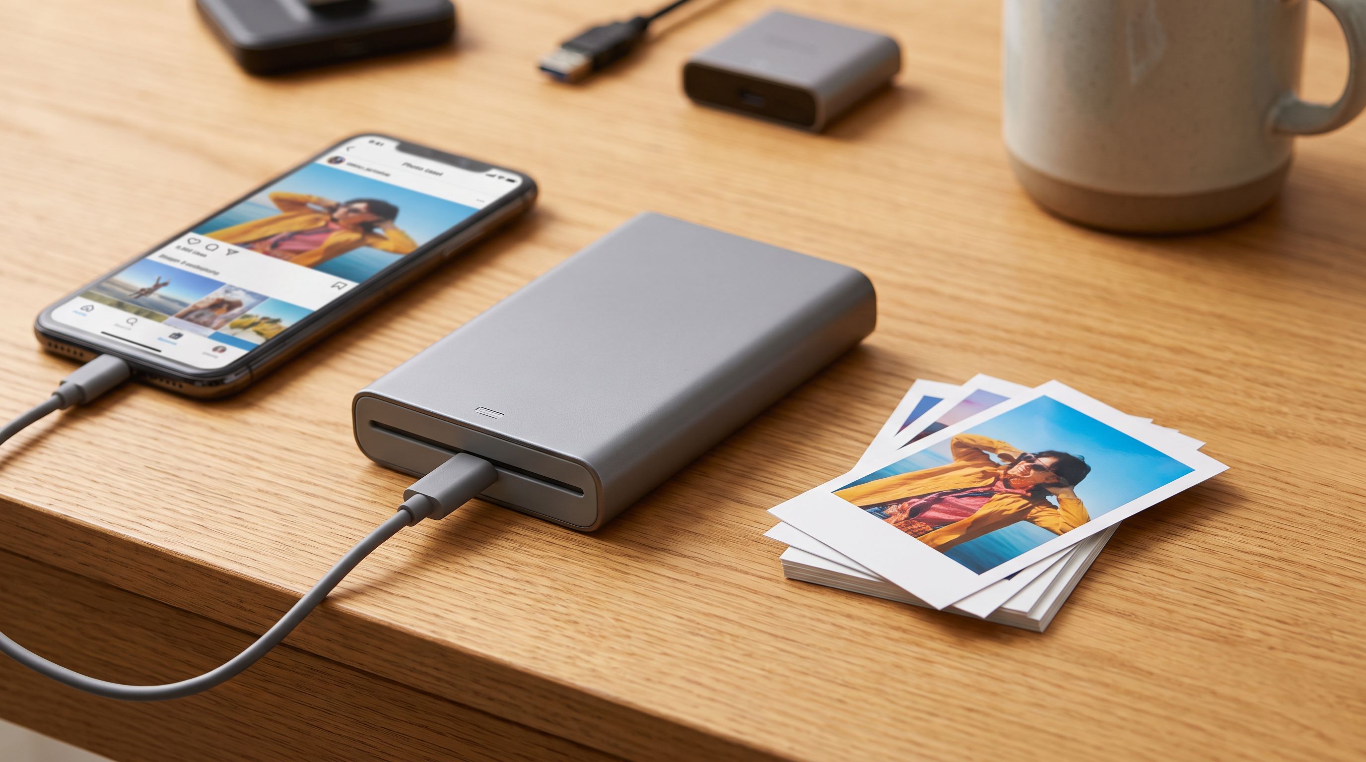 A pocket-sized portable photo printer beside a smartphone and a stack of freshly printed 2x3 inch photos on a wooden desk