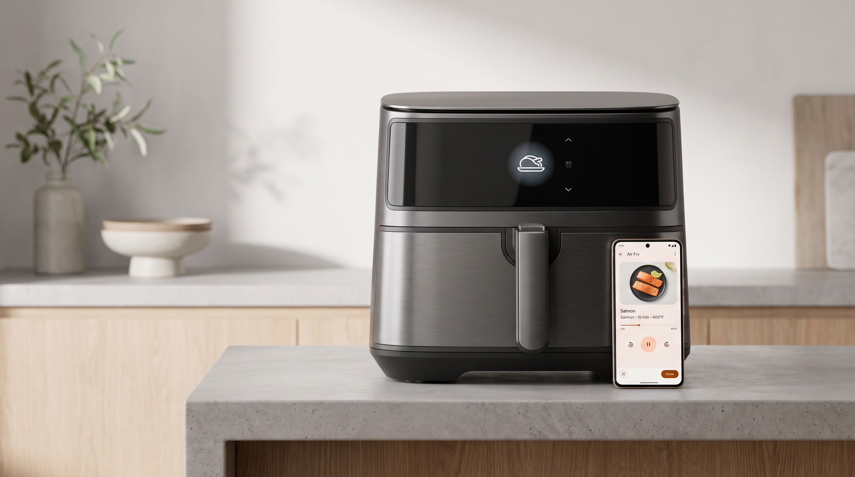 A modern smart air fryer on a kitchen counter with a smartphone app showing cooking controls beside it