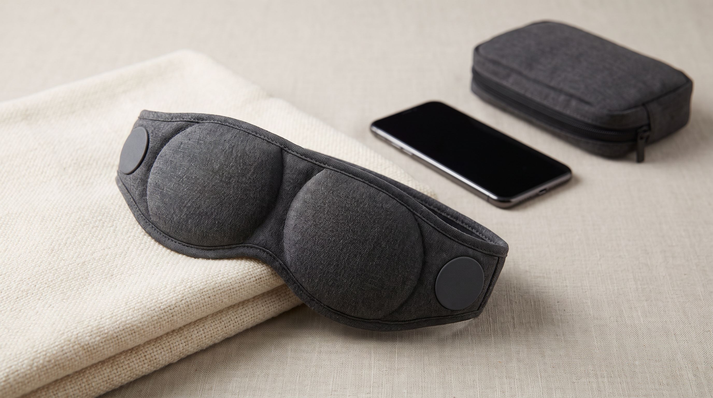 A padded contoured sleep mask with built-in flat speakers resting on a folded blanket next to a phone and travel pouch