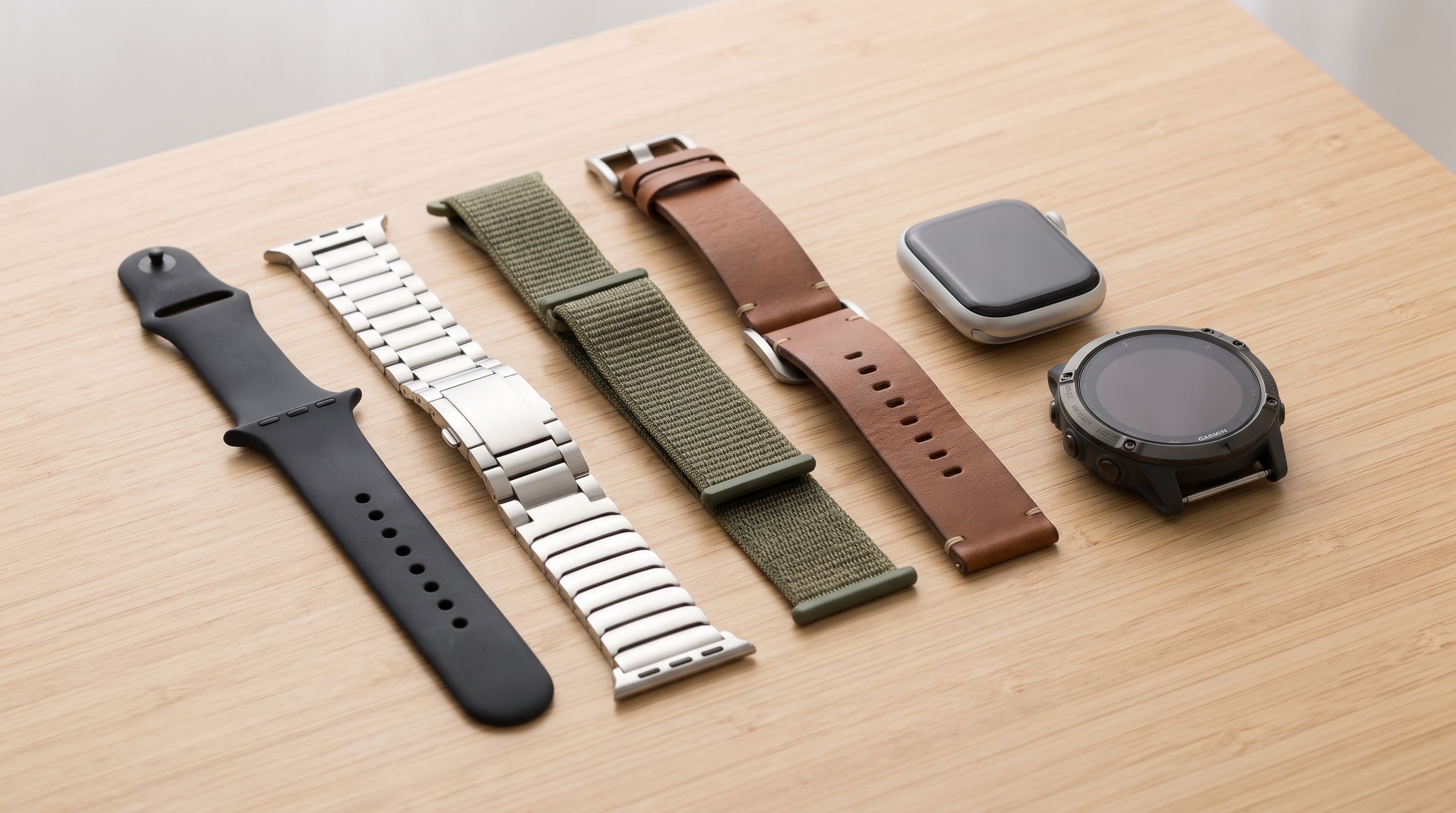 Several smartwatch bands laid flat on a wooden surface — a silicone sport band, a stainless steel link bracelet, a woven nylon loop and a leather strap — beside an Apple Watch and a Garmin watch body