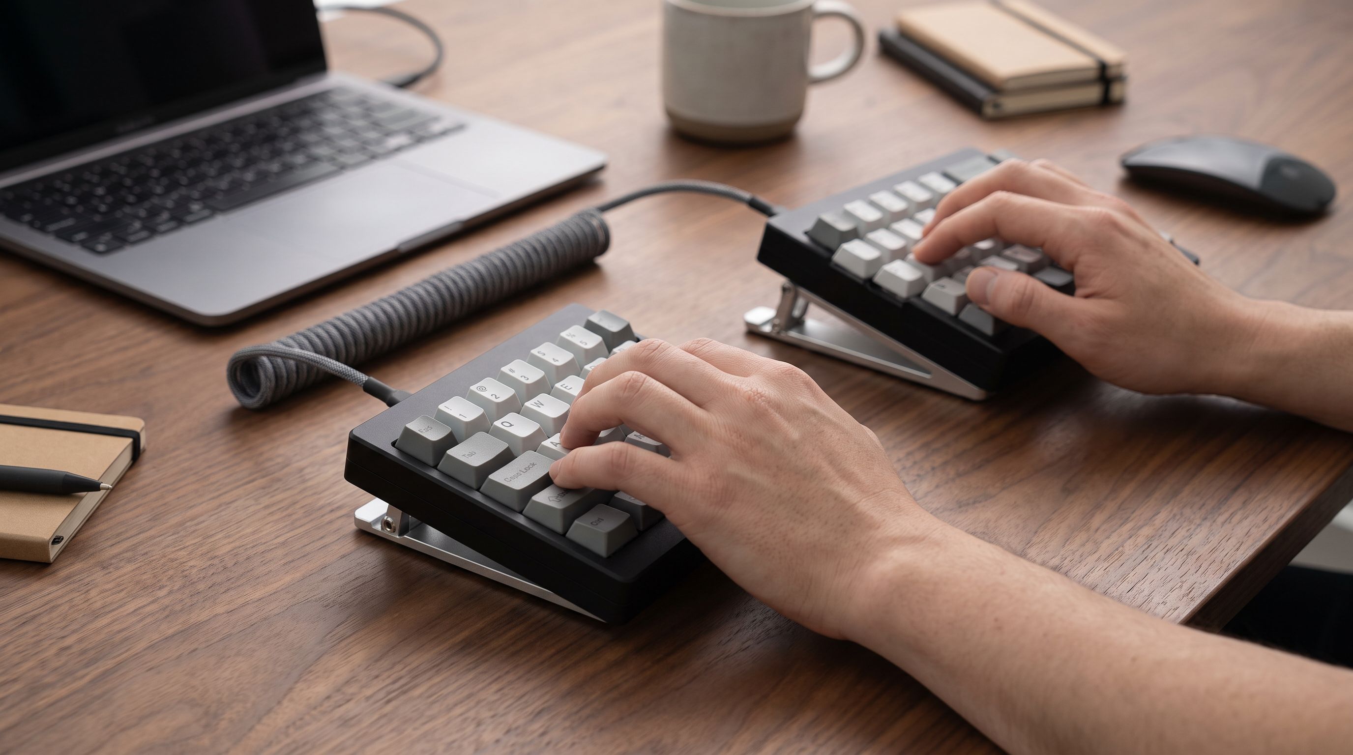 A split mechanical keyboard with both halves separated and tented at an angle on a wooden desk, with a person's hands resting in a neutral, shoulder-width typing position