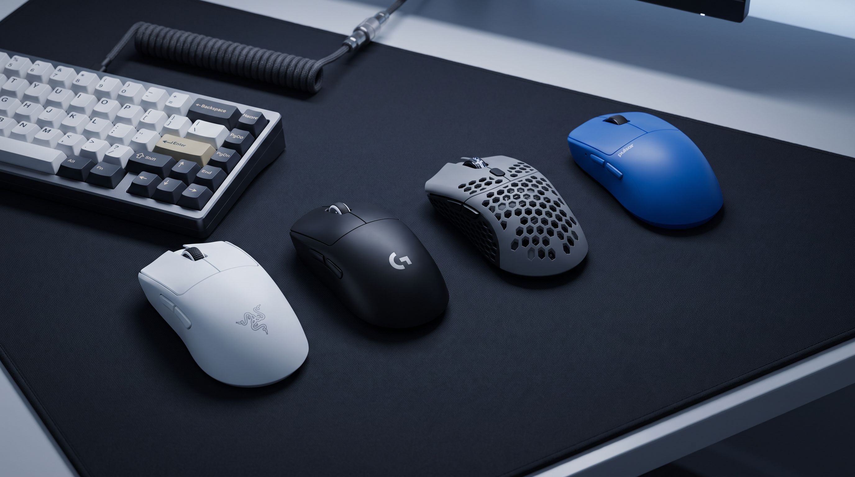 Four lightweight wireless gaming mice arranged on a large cloth mousepad next to a mechanical keyboard, lit by cool desk lighting
