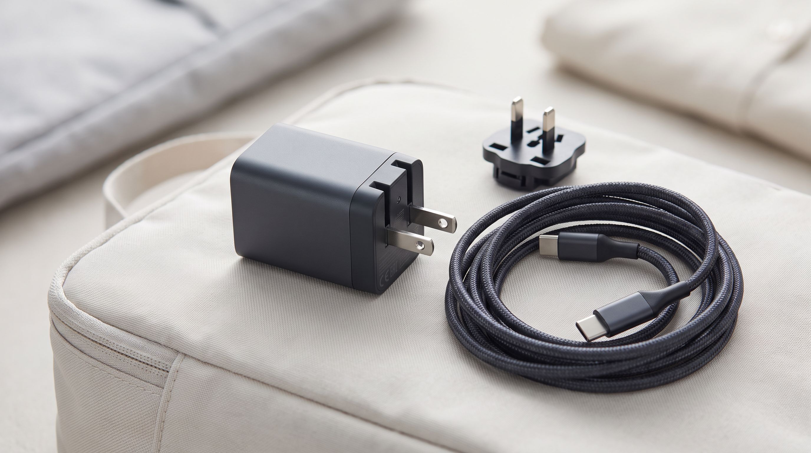 A compact GaN USB-C travel charger with folding prongs beside a braided USB-C cable and international plug adapter on a packing cube