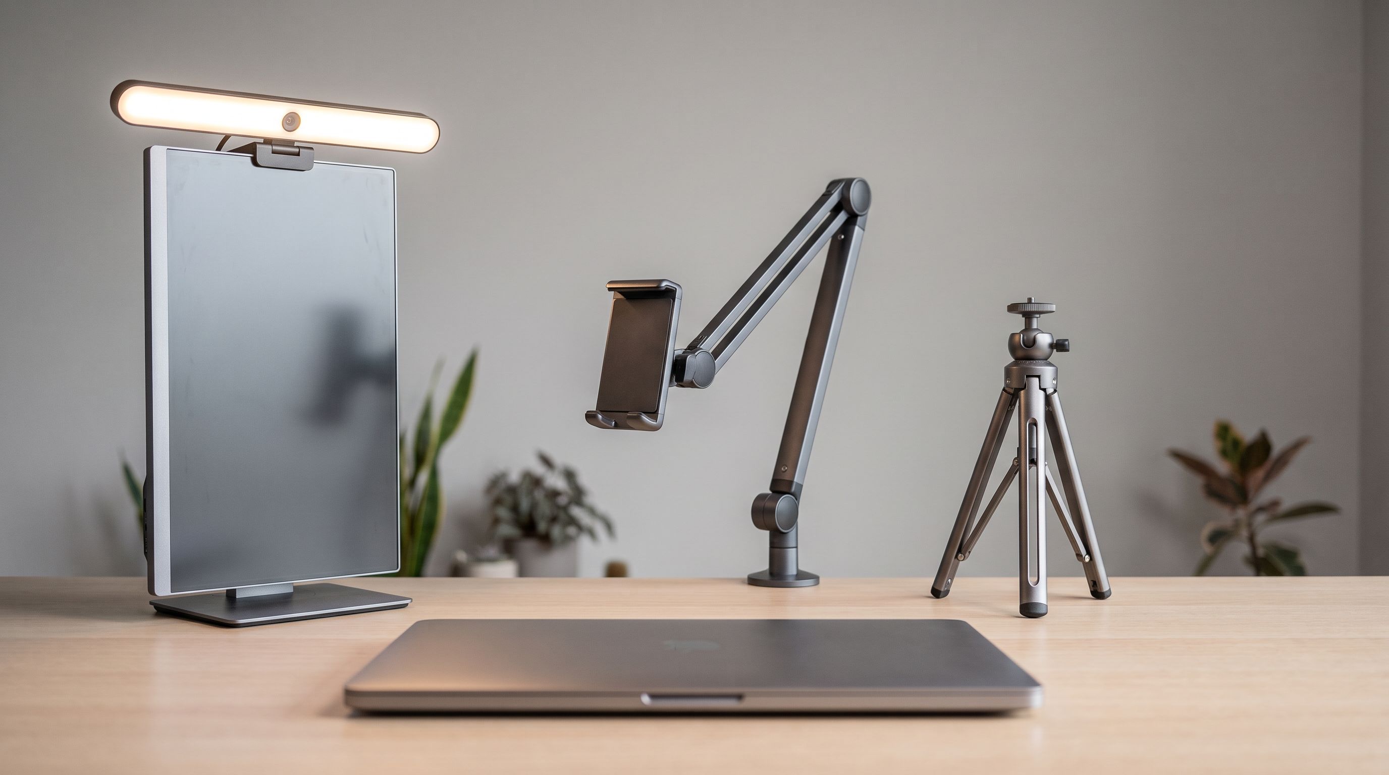 A desk setup showing a webcam light on a monitor clip, an articulating desk arm, and a small tripod side by side behind a laptop