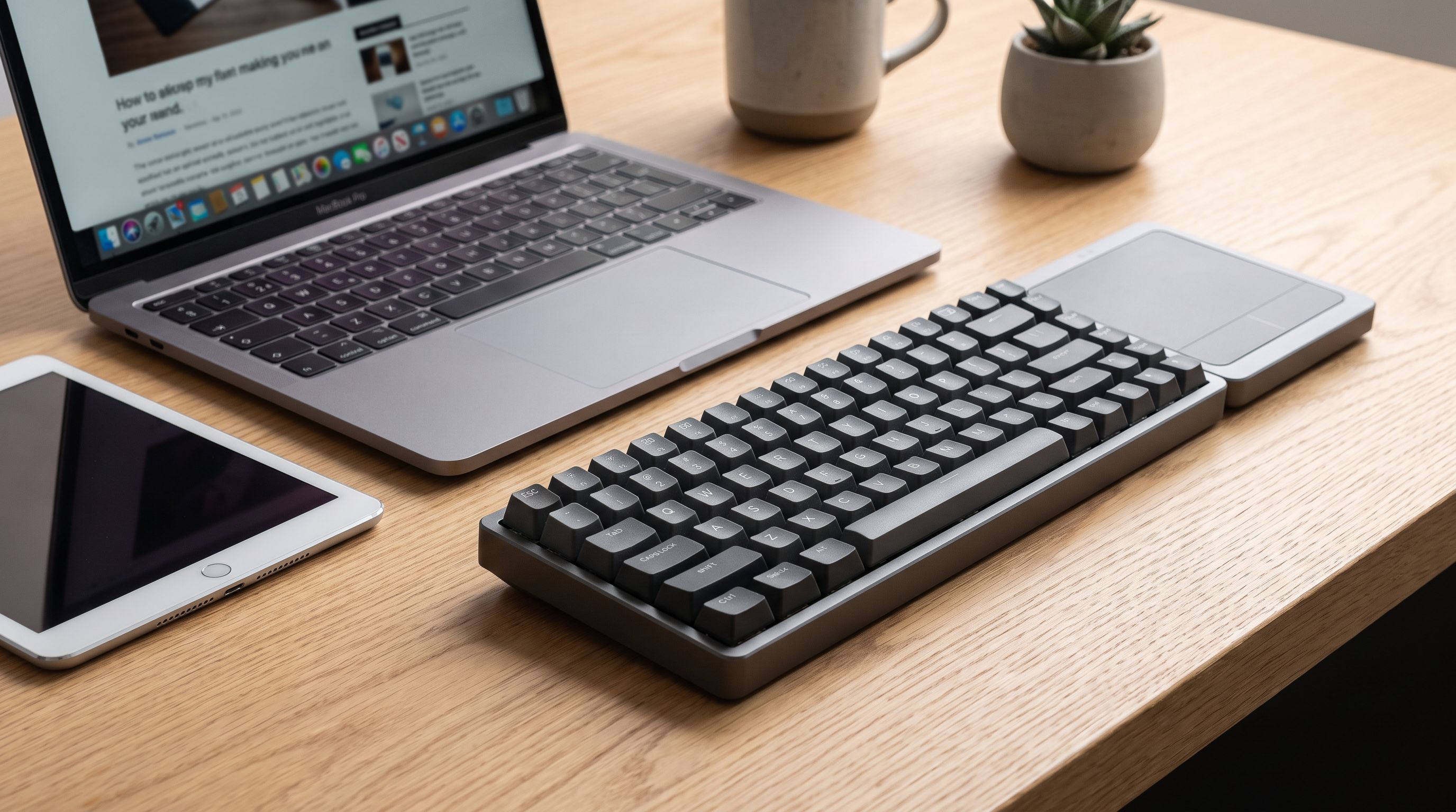 A low-profile wireless keyboard and trackpad on a wooden desk beside a MacBook and an iPad