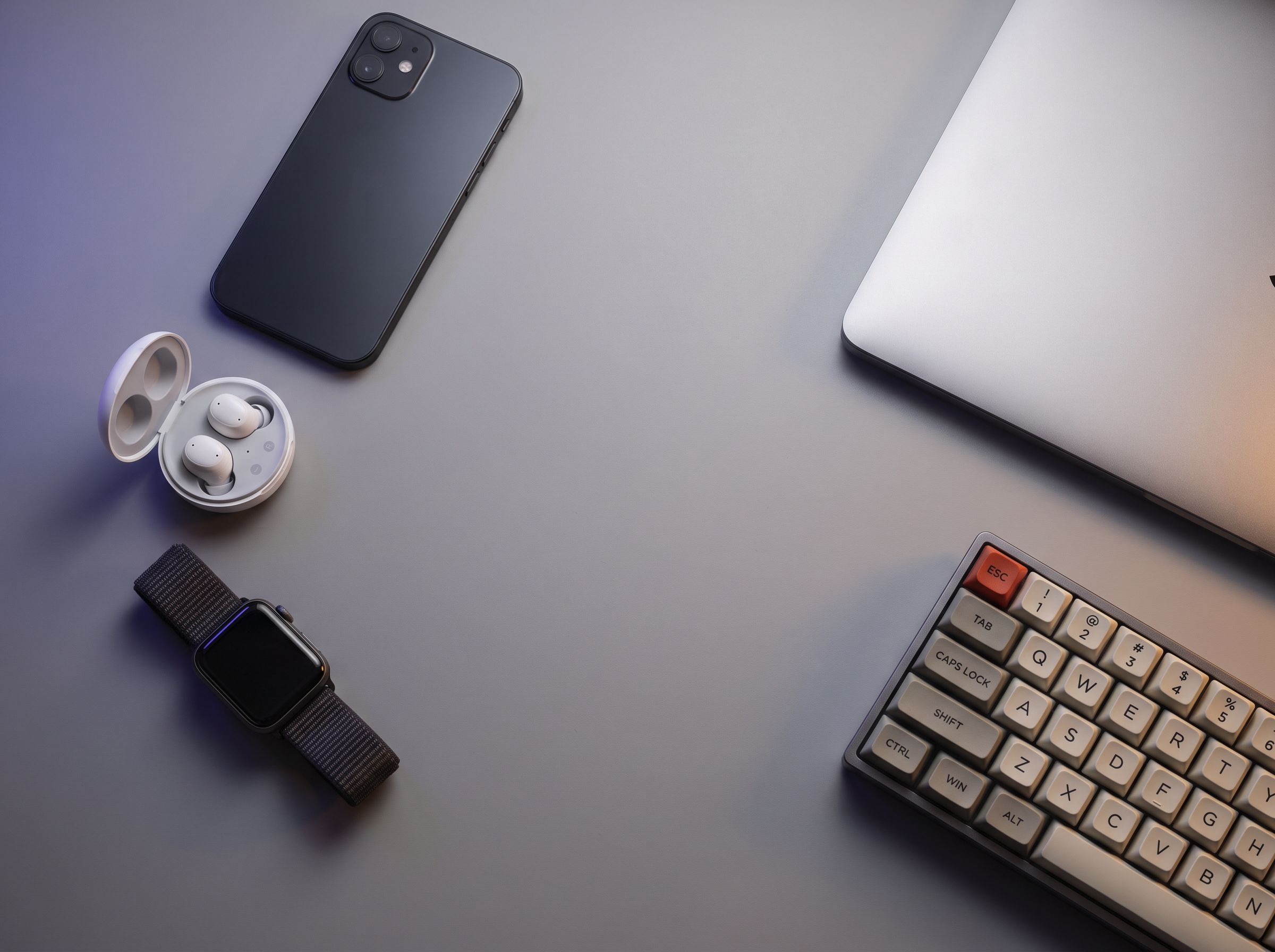 A modern desk flat-lay of tech gadgets: a smartphone, wireless earbuds, a smartwatch, a laptop and a mechanical keyboard